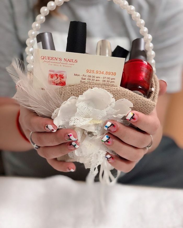 Hands with colorful nail art holding a small pearl-handled gift basket with polish and a Queen's Nails business card